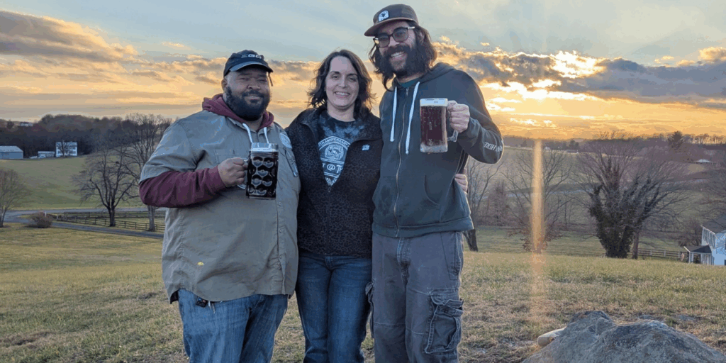 2 men holding beer with a woman in between standing in an open farm field