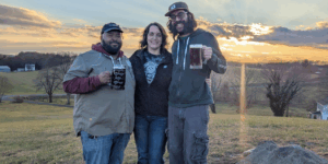 2 men holding beer with a woman in between standing in an open farm field