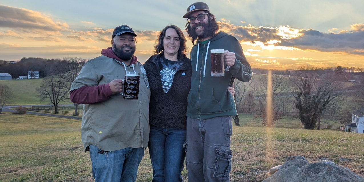 2 men holding beer with a woman in between standing in an open farm field